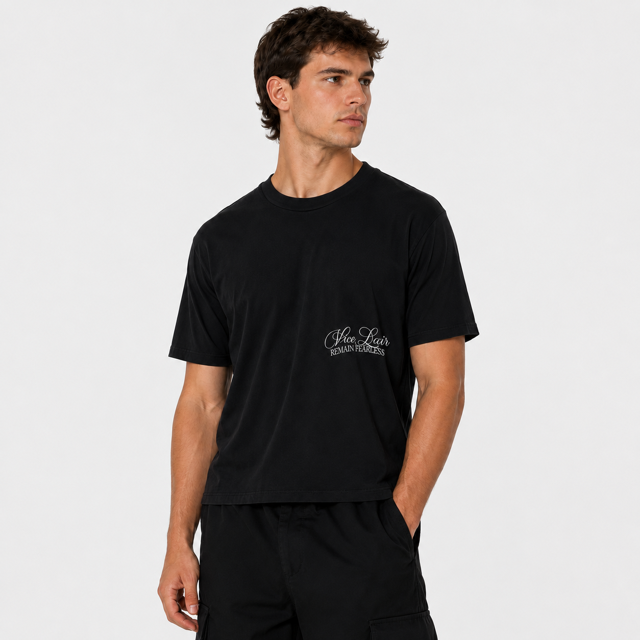 A man wearing a black short-sleeve crewneck t-shirt with a small white script logo on the left chest, paired with black pants, against a light gray studio background.