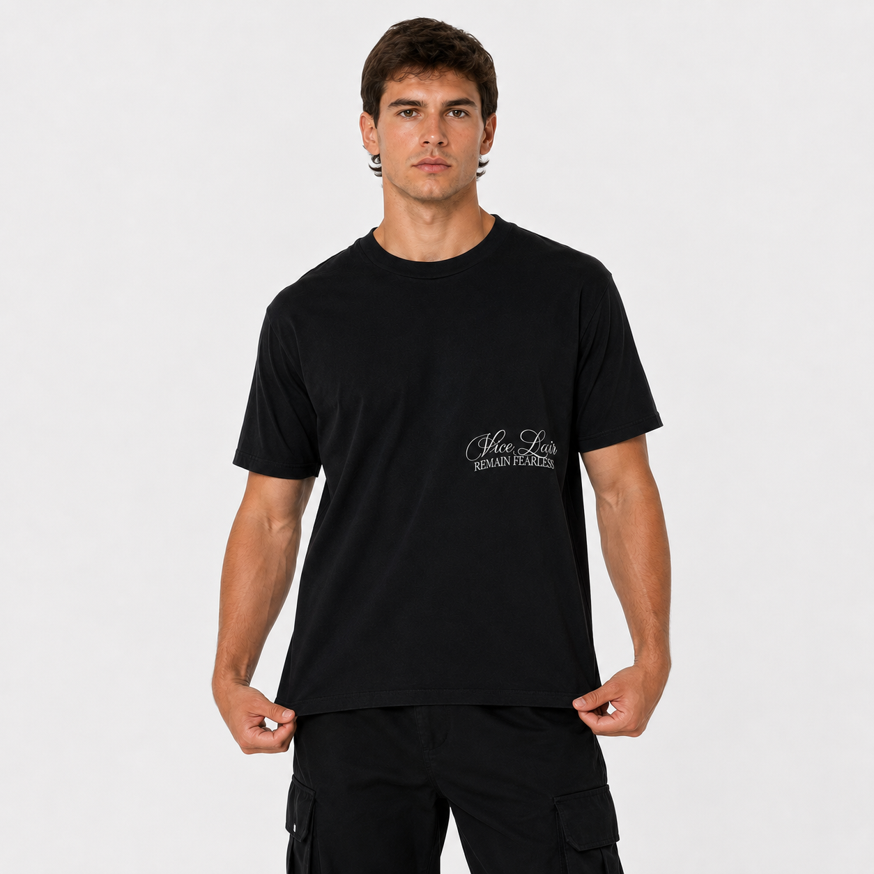 A man wearing a black short-sleeve crewneck t-shirt with a small white script logo on the left chest, paired with black cargo pants, against a light gray studio background.
